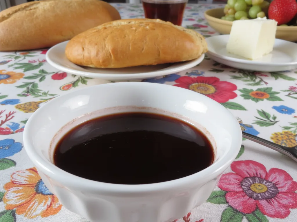 Kahvaltı sofrasında beyaz kasede üzüm pekmezi; arkada ekmek, beyaz peynir ve çiçekli masa örtüsü