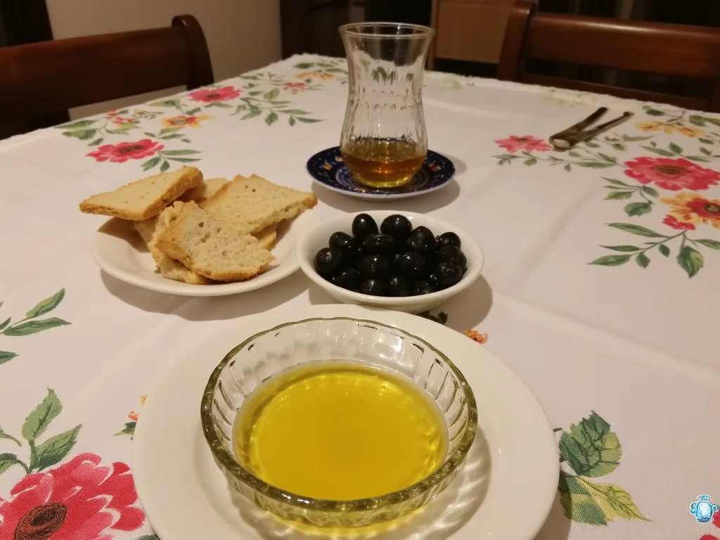 Çiçekli örtülü sofrada cam kasede sızma zeytinyağı; yanında ekmek parçaları, bir kase siyah zeytin ve çay bardağı